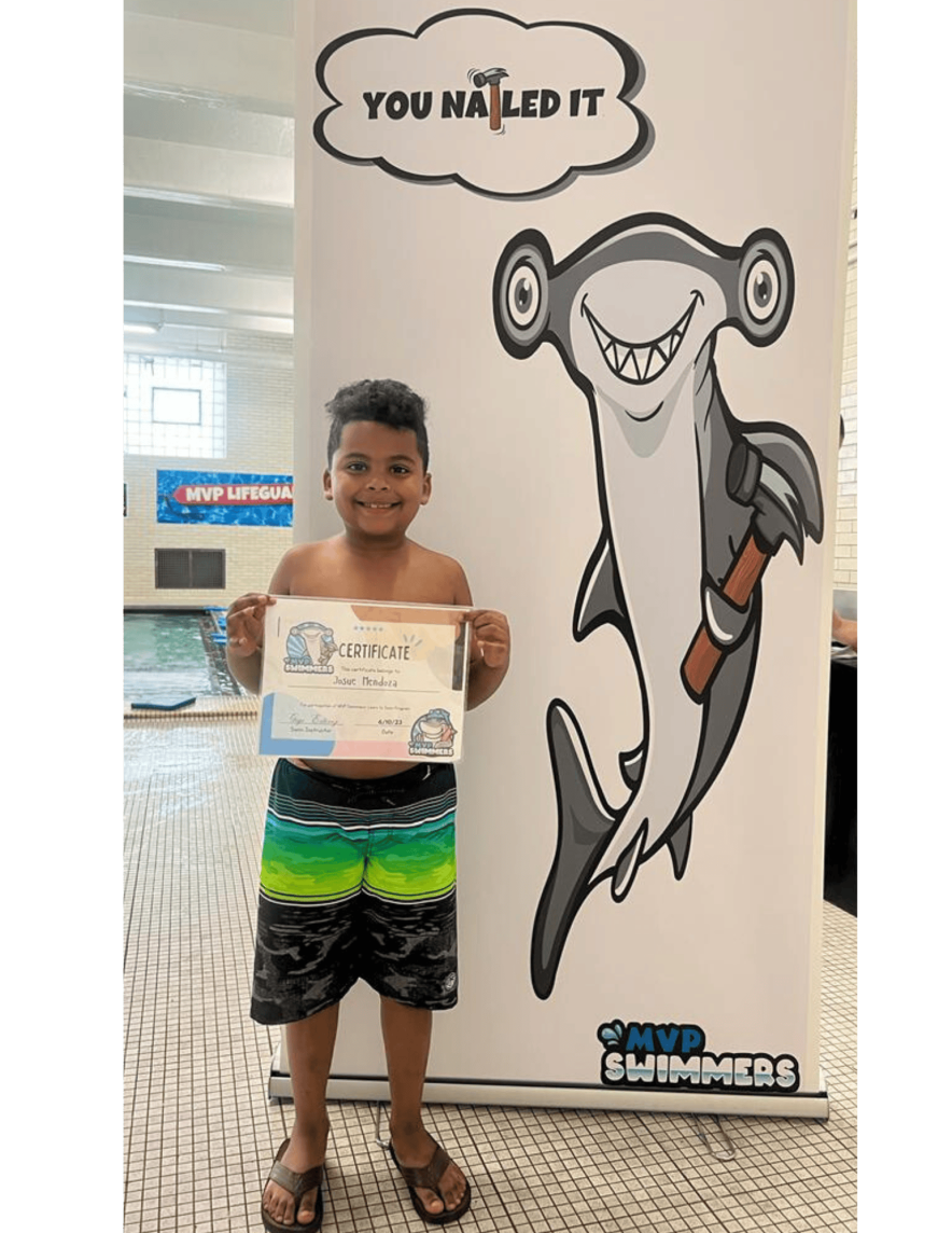 A young swimmer holding his certificate beside the MVP Swimmers "You nailed it" banner